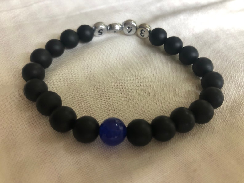 May include: Black beaded bracelet with a single blue bead and silver number beads. The bracelet is made of matte black beads and has a stretchable design. The numbers on the silver beads are 3, 4, and 5.