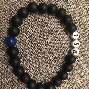 May include: Black beaded bracelet with a single blue bead and white beads with the numbers 1, 5, and 6. The bracelet is made of matte black beads. The bracelet is on a gray textured surface.