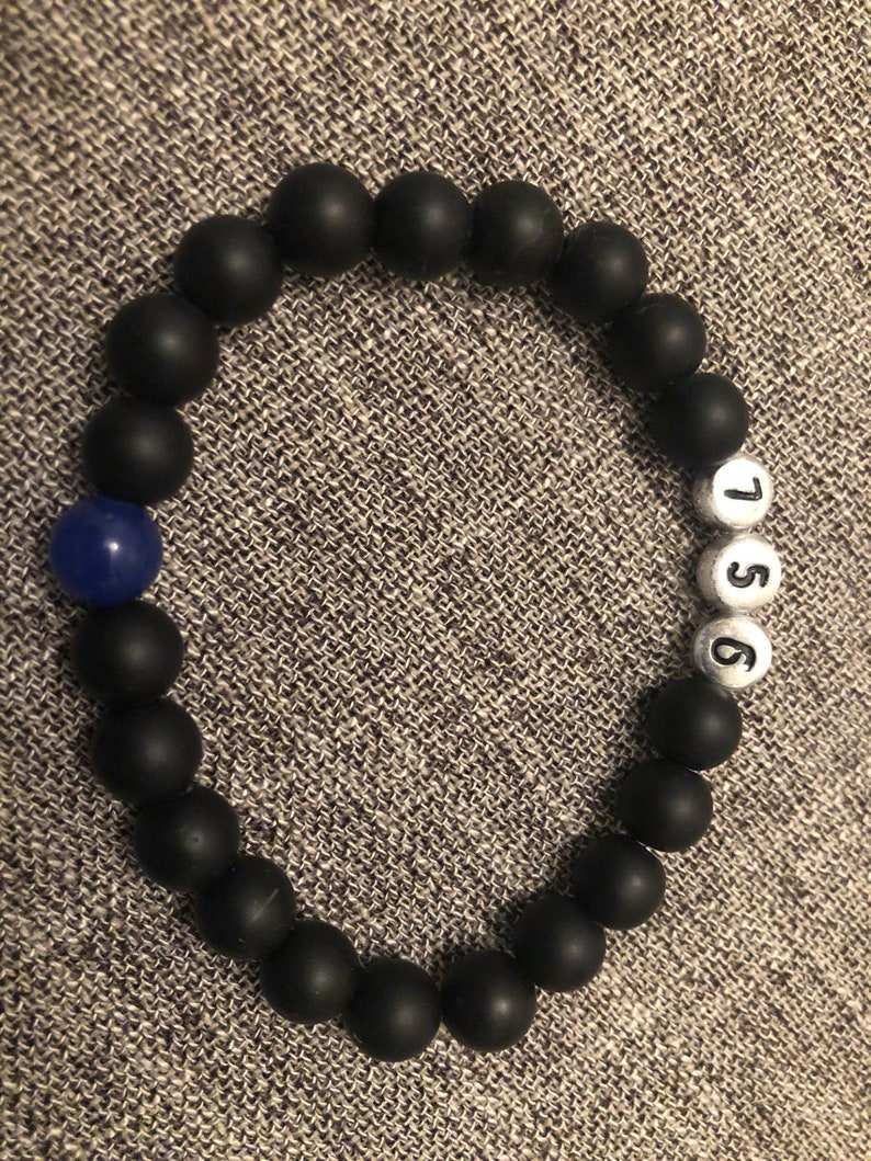 May include: Black beaded bracelet with a single blue bead and white beads with the numbers 1, 5, and 6. The bracelet is made of matte black beads. The bracelet is on a gray textured surface.