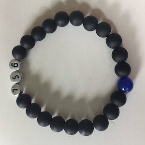 May include: Black beaded bracelet with silver number beads and a single blue bead. The bracelet features matte black round beads and silver beads with the numbers 5, 6, and 7. A single, glossy blue bead adds a pop of color.