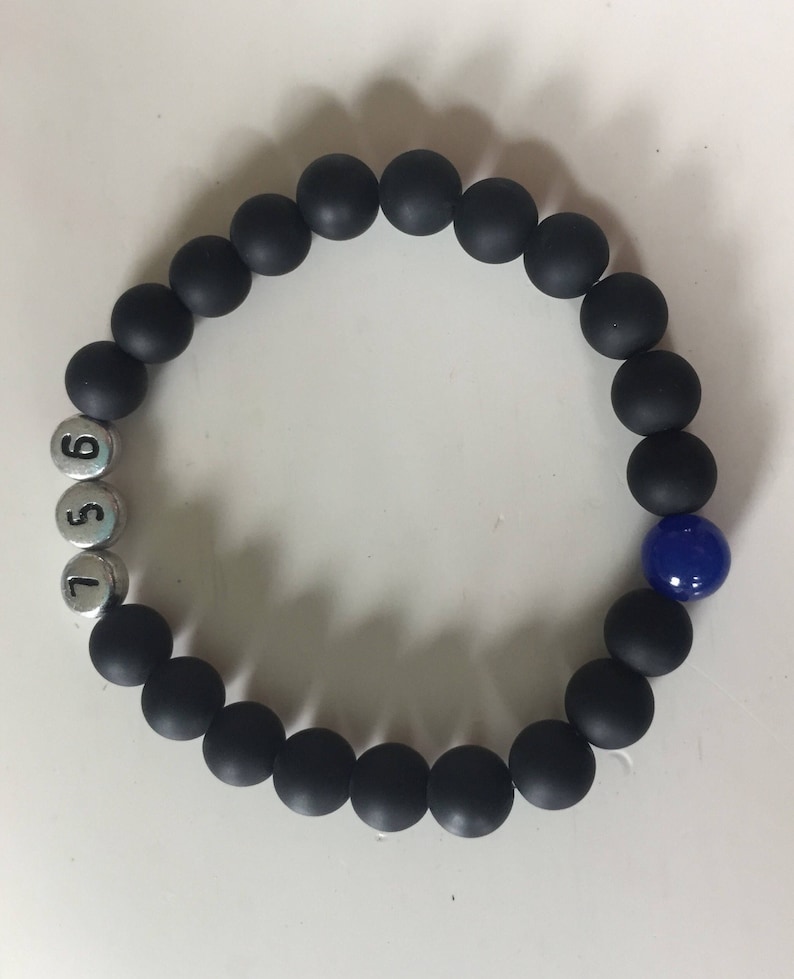 May include: Black beaded bracelet with silver number beads and a single blue bead. The bracelet features matte black round beads and silver beads with the numbers 5, 6, and 7. A single, glossy blue bead adds a pop of color.