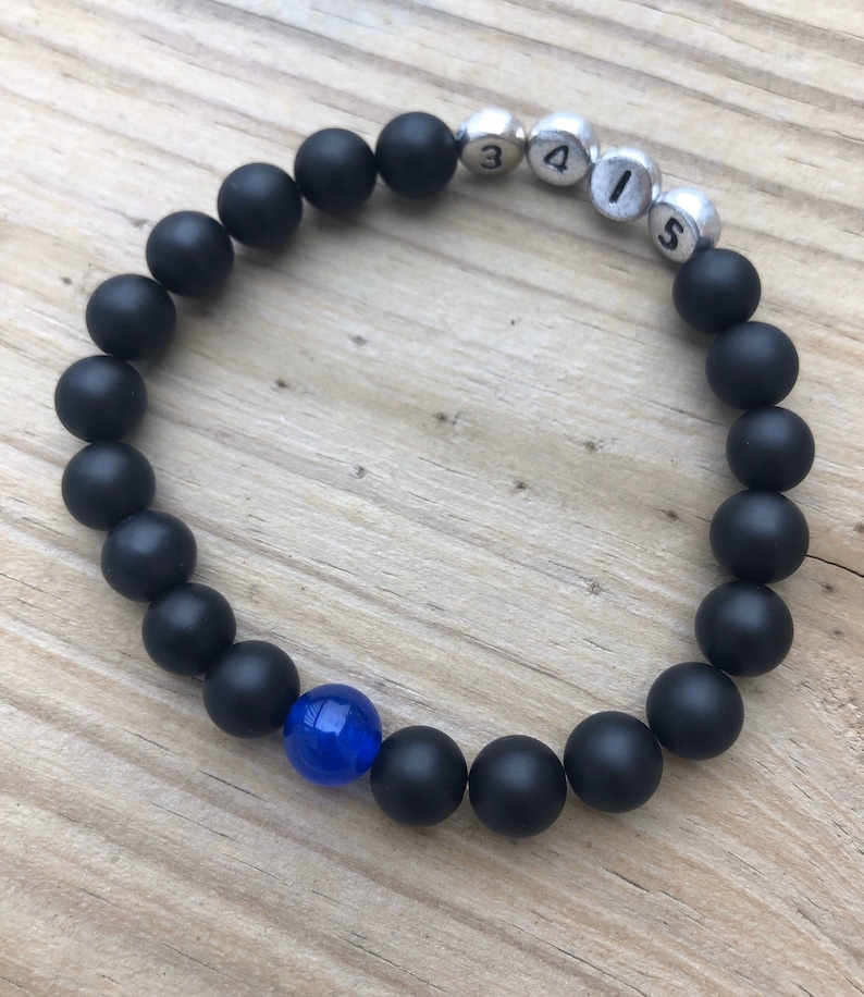 May include: A black beaded bracelet with a single blue bead and silver beads with the numbers "3", "4", "1", and "5". The bracelet is on a wooden surface.