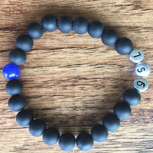 May include: A black beaded bracelet with a single blue bead and three silver beads with the numbers 1, 5, and 6. The bracelet is displayed on a wooden surface.