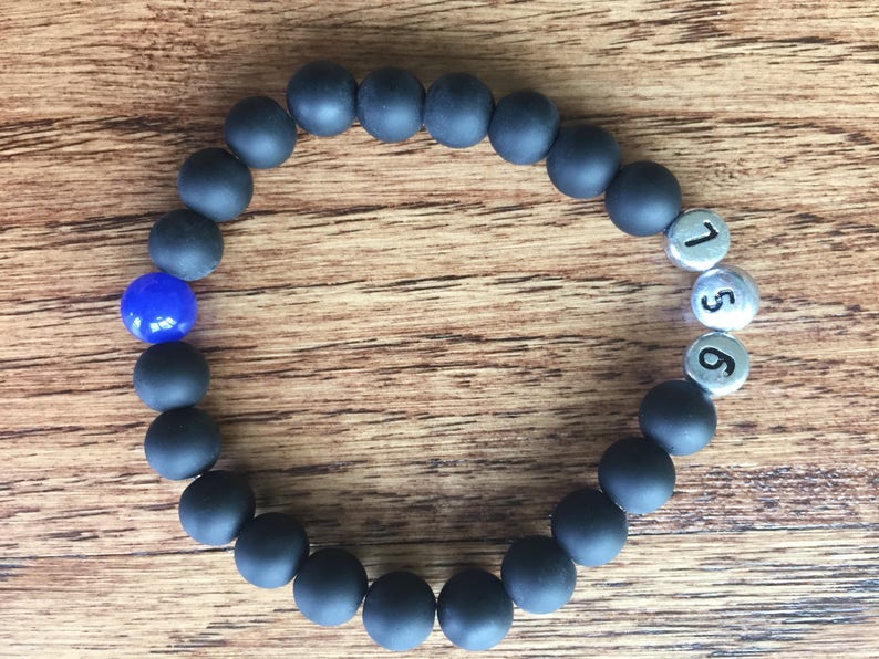 May include: A black beaded bracelet with a single blue bead and three silver beads with the numbers 1, 5, and 6. The bracelet is displayed on a wooden surface.