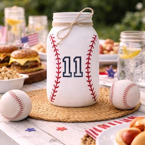 May include: A white mason jar decorated as a baseball, with red stitching and the number 11 in navy blue. Two baseballs and food items are on a wooden table, suggesting a baseball-themed party.