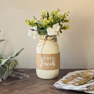 May include: White painted mason jar with a burlap ribbon that says "God Bless Jacob" and a bouquet of white flowers with green leaves.