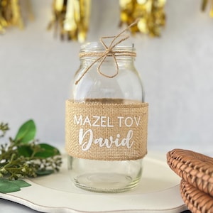 May include: A clear glass jar with a burlap band that says "Mazel Tov David" in white lettering. The jar is tied with twine and is sitting on a white surface.