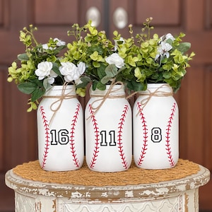 May include: Three white mason jars with red stitching and black numbers, 16, 11, and 8, are filled with green and white floral arrangements. The jars are sitting on a white table with a brown mat.
