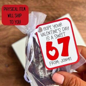 May include: A small gift bag tied with white ribbon, holding a product. A red tag with the text "Hope your Valentine's Day is a sweet 67" and "From: Jonah" is attached. The tag has a white heart and red numbers.