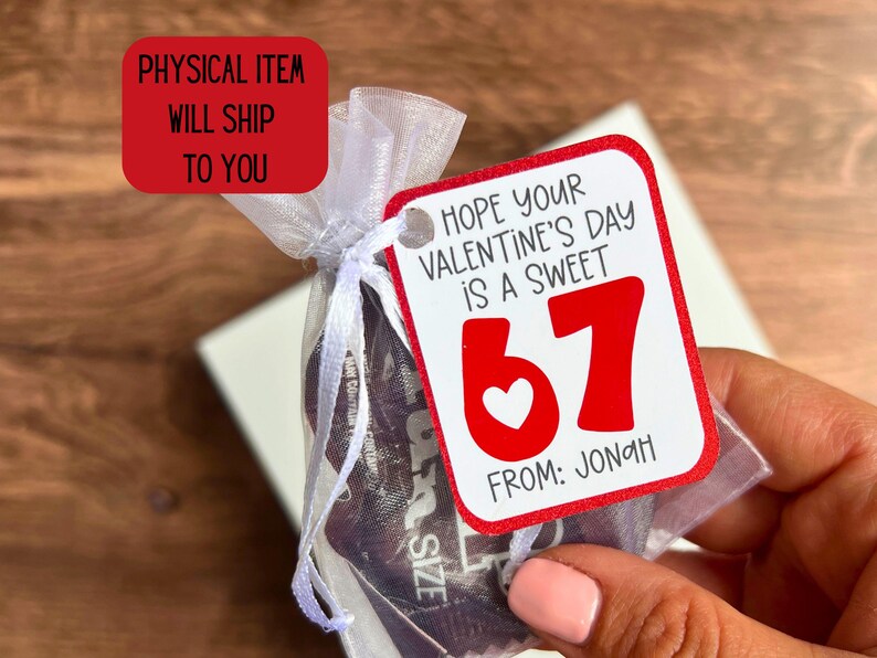 May include: A small gift bag tied with white ribbon, holding a product. A red tag with the text "Hope your Valentine's Day is a sweet 67" and "From: Jonah" is attached. The tag has a white heart and red numbers.