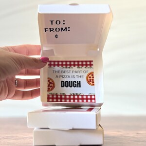 May include: A white pizza box with a red and white checkered pattern and the text "The best part of a pizza is the dough" with a small pizza graphic. The box is open and the top is folded back.