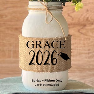 May include: A white painted mason jar with a burlap band that reads "GRACE 2026" with a graduation cap graphic. The jar is tied with twine and contains greenery. The text "Burlap + Ribbon Only Jar Not Included" is at the bottom.