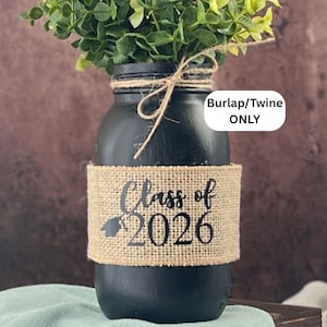 May include: A black painted mason jar with a burlap band that reads "Class of 2026". The jar is tied with twine and filled with greenery. A white oval sticker on the right says "Burlap/Twine ONLY".