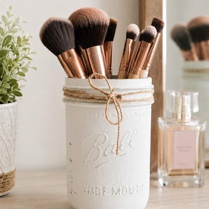 May include: A white painted glass jar with the embossed text Ball and WIDE MOUTH, wrapped with a thin jute twine bow. The jar contains several makeup brushes with copper-colored metal ferrules and soft brown and black bristles.