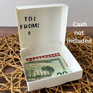 May include: White cardboard pizza box with a red and white checkered pattern inside. The box has a blank label with the words "TO:" and "FROM:" written on it. The text "Cash not included" is written on the side of the box.