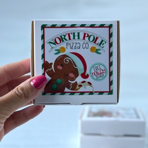 May include: A white cardboard pizza box with a green and white striped border. The box features a cartoon gingerbread person holding a slice of pizza with the text "North Pole Pizza Co." and "Free Holiday Delivery".