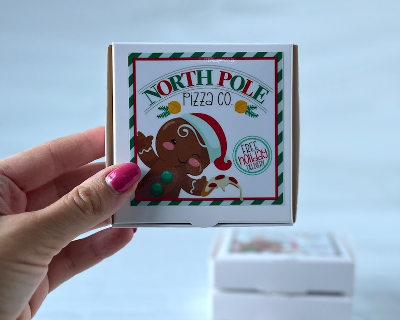 May include: A white cardboard pizza box with a green and white striped border. The box features a cartoon gingerbread person holding a slice of pizza with the text "North Pole Pizza Co." and "Free Holiday Delivery".
