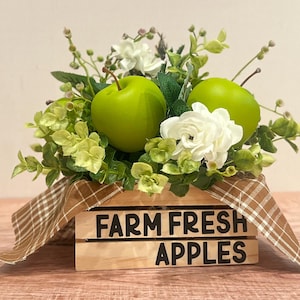 May include: A wooden crate displaying "FARM FRESH APPLES" contains green apples, white flowers, and foliage. A brown plaid ribbon adorns the crate, giving a rustic, farmhouse appearance.