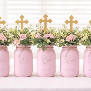 May include: Five pink mason jars filled with flowers and topped with gold crosses. Each jar is adorned with a twine bow. The jars are arranged on a white surface, creating a decorative centerpiece.
