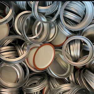 May include: A close-up shot of a pile of canning lids and rings. The lids are silver-colored with a white interior and a red-orange rubber seal. The rings are also silver and have a textured design.