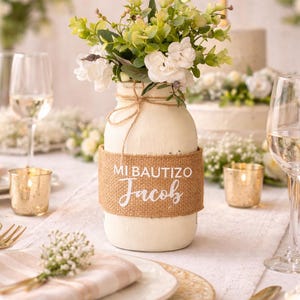 May include: A white painted jar with a burlap band that reads "Mi Bautizo Jacob" holds a floral arrangement of white and green flowers. The jar is decorated with twine. The table setting includes gold cutlery, candles, and wine glasses.