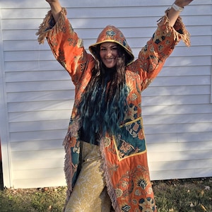 May include: An orange, floral-patterned coat with a hood and fringe trim. The coat is paired with yellow floral trousers. The coat has a teal and gold pocket. The person is smiling and has their arms raised in a peace sign.