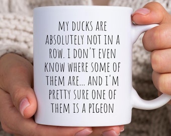 May include: White ceramic coffee mug with black text that reads "My ducks are absolutely not in a row. I don't even know where some of them are... and I'm pretty sure one of them is a pigeon."