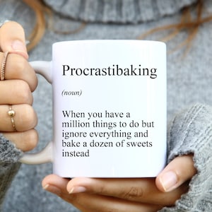 May include: White ceramic mug with the text "Procrastibaking (noun) When you have a million things to do but ignore everything and bake a dozen of sweets instead."