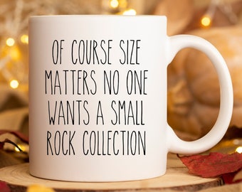 May include: White ceramic mug with black text that reads "Of course size matters no one wants a small rock collection". The mug is sitting on a wooden surface with autumn leaves and fairy lights in the background.