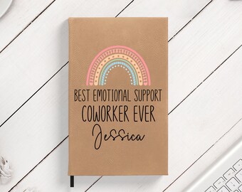 May include: A brown leather-bound journal with a rainbow design and the text "Best Emotional Support Coworker Ever" and the name "Jessica" written in cursive.