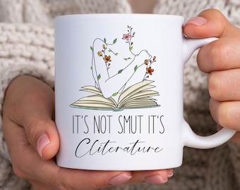 May include: White ceramic mug with a black and white illustration of a person reading a book with flowers growing from their body. The text on the mug reads "It's not smut it's Cliterature".