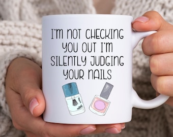 Op de afbeelding: Witte keramische mok met een zwart-wit ontwerp. De mok heeft de tekst "I'm not checking you out I'm silently judging your nails" en twee illustraties van nagellak flesjes.