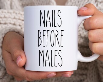 Op de afbeelding: Witte keramische mok met de tekst "NAILS BEFORE MALES" in zwart, vastgehouden door een persoon die een crèmekleurige trui draagt. De mok heeft een standaard handvat en een glanzende afwerking.