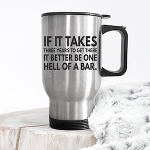 May include: Stainless steel travel mug with a black lid and handle. The mug has a black graphic with the text "If it takes three years to get there it better be one hell of a bar."