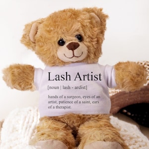 Op de afbeelding: Een bruine teddybeer die een wit T-shirt draagt met de tekst "Lash Artist [noun | lash - ardist] hands of a surgeon, eyes of an artist, patience of a saint, ears of a therapist."