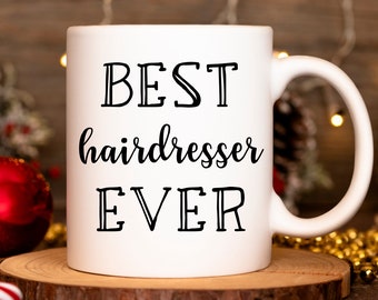 Peut inclure: Une tasse en céramique blanche avec du texte noir qui dit "BEST hairdresser EVER". La tasse est posée sur un dessous de tasse en bois.