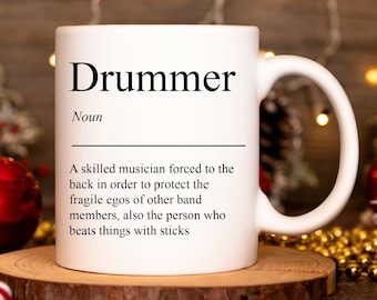 Op de afbeelding: Een witte keramische mok met een zwart-witte definitie van het woord "Drummer". De definitie luidt: "Een bekwame muzikant die gedwongen wordt om achterin te staan om de kwetsbare ego's van andere bandleden te beschermen, ook de persoon die dingen met stokken slaat."