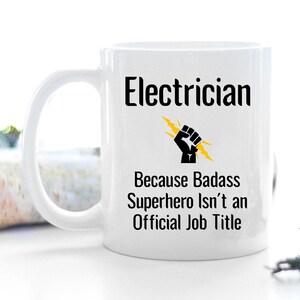 May include: White ceramic mug with the word "Electrician" in black, above the text "Because Badass Superhero Isn't an Official Job Title." A black fist with yellow lightning bolts is in the center.