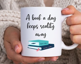 May include: White ceramic mug with a black and blue illustration of a stack of books and the text "A book a day keeps reality away".