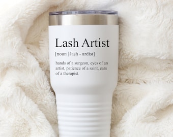 Puede incluir: Un vaso térmico blanco con borde y tapa plateados. El texto "Lash Artist" está impreso en la parte delantera, junto con una definición. El vaso es cilíndrico con un diseño acanalado y está sobre un fondo suave, blanco y texturizado.