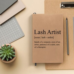 May include: A tan leather-bound journal with the text "Lash Artist" and the definition "[noun | lash - ardist] hands of a surgeon, eyes of an artist, patience of a saint, ears of a therapist."
