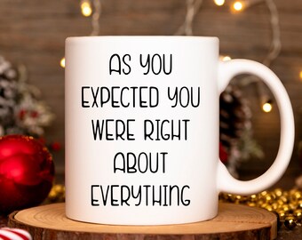 May include: White ceramic coffee mug with black text that reads "As you expected you were right about everything". The mug is sitting on a wooden surface with a red bauble and gold beads in the background.