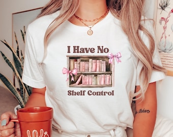 May include: White t-shirt with the text "I Have No Shelf Control" and a graphic of a bookshelf with pink books and bows. A terracotta mug with cactus designs is held in the hand. The person is wearing a gold scrunchie and jewelry.