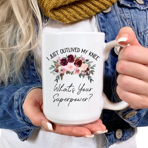 Pu&ograve; includere: Una tazza in ceramica bianca con un motivo floreale e il testo "I just outlived my knee. What's your superpower?"