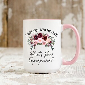 Pu&ograve; includere: Una tazza in ceramica bianca con bordo e manico rosa. La tazza presenta un motivo floreale con fiori color borgogna e rosa e il testo "I just outlived my knee. What's your superpower?"
