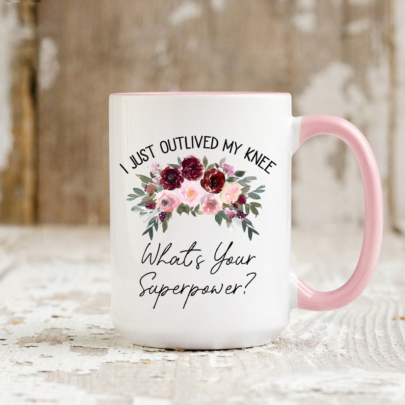 Pu&ograve; includere: Una tazza in ceramica bianca con bordo e manico rosa. La tazza presenta un motivo floreale con fiori color borgogna e rosa e il testo "I just outlived my knee. What's your superpower?"