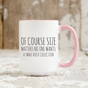 May include: White ceramic coffee mug with a pink rim and handle. The mug has black text that reads "Of course size matters no one wants a small rock collection."