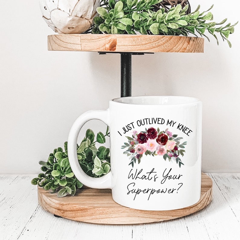 Pu&ograve; includere: Una tazza da caff&egrave; in ceramica bianca con un motivo floreale nero e il testo "I just outlived my knee. What's your superpower?"