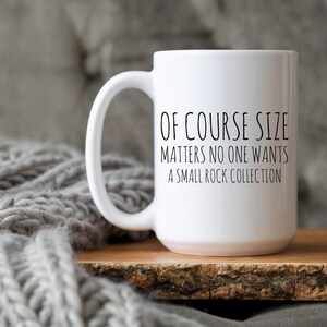 May include: White ceramic coffee mug with black text that reads "Of course size matters no one wants a small rock collection". The mug is sitting on a wooden surface.