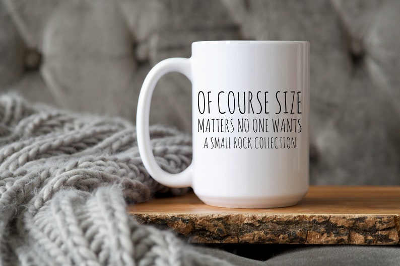 May include: White ceramic coffee mug with black text that reads "Of course size matters no one wants a small rock collection". The mug is sitting on a wooden surface.